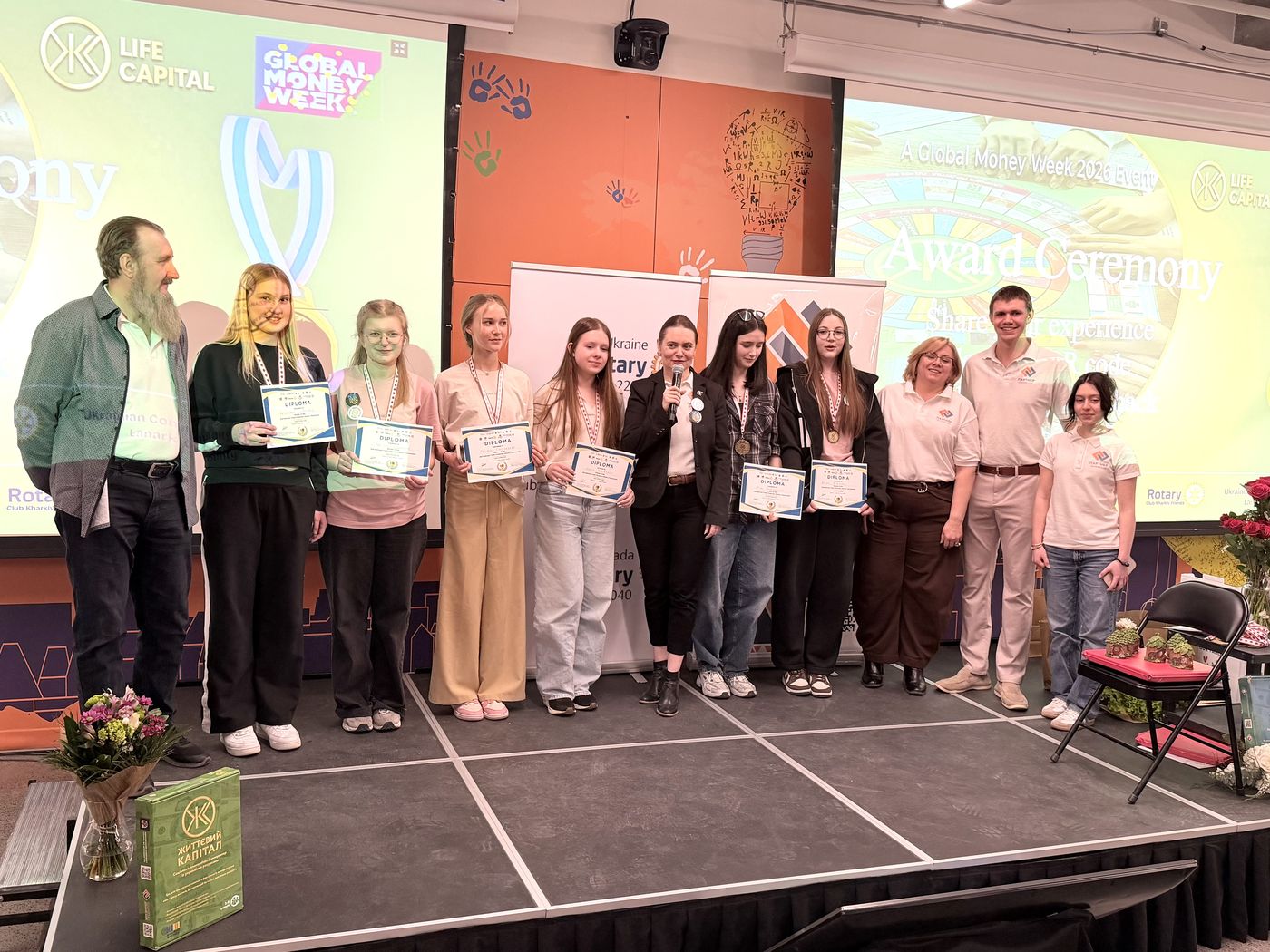 Six participants of the 2nd National Youth Financial Literacy Tournament hold their diplomas on stage at the award ceremony in Ottawa, 14 March 2026, with the organisers beside them.